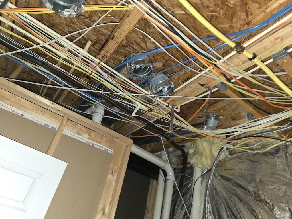 Numerous colorful wires, pipes, and ducts crisscross an unfinished wooden ceiling. TITLE