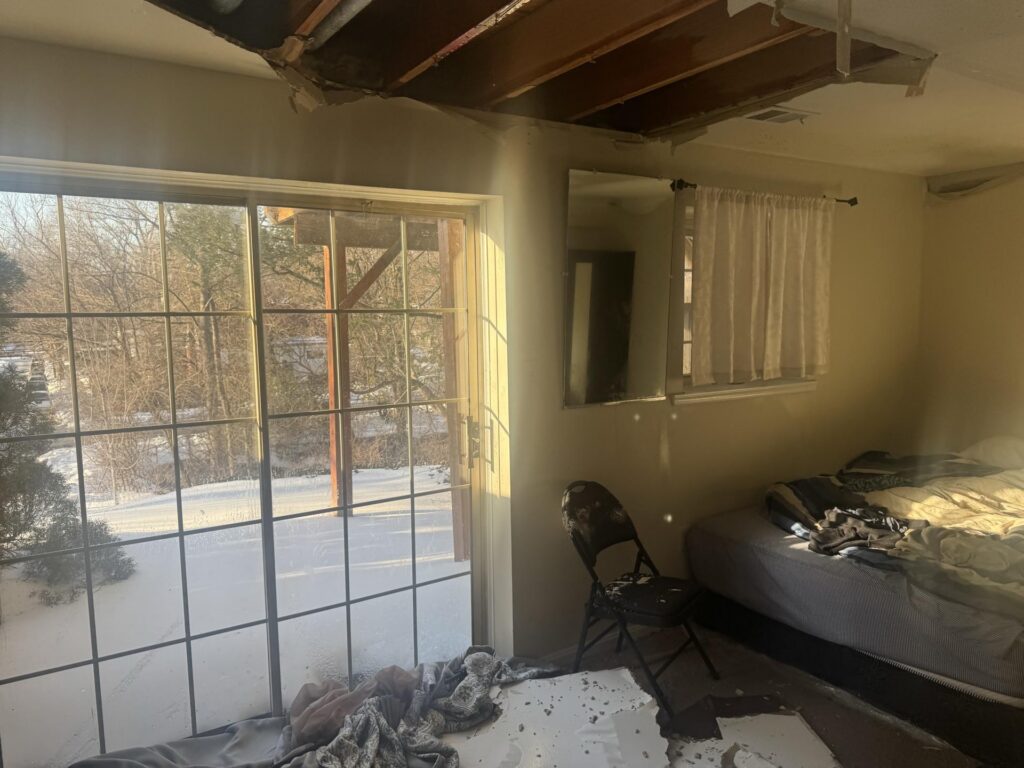 Sunlight streams into a room with a damaged ceiling and debris, overlooking a snowy winter scene.