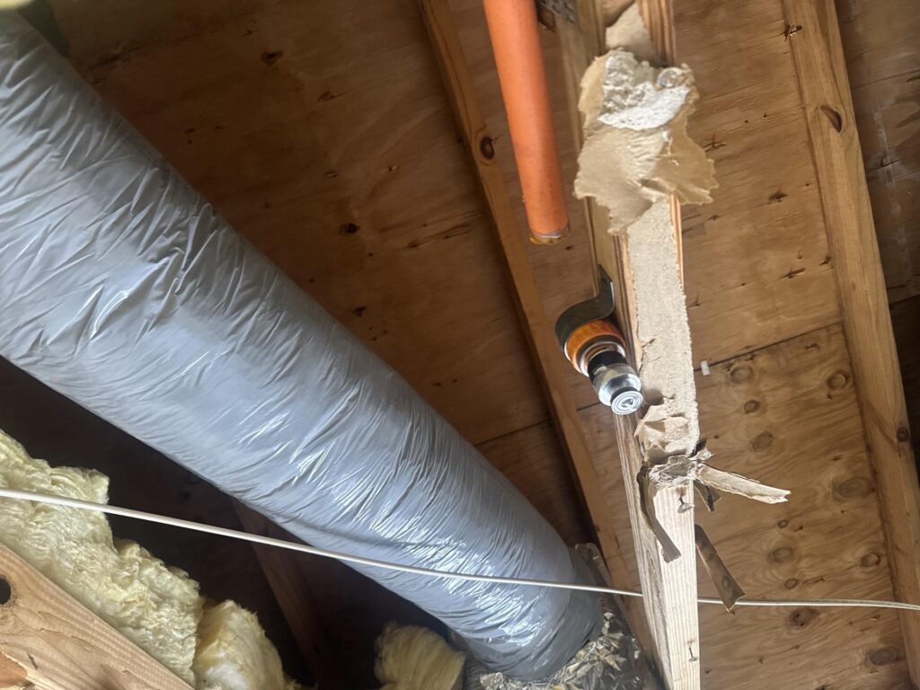 An attic interior features a large silver HVAC duct, orange pipe, wooden beams, and insulation.