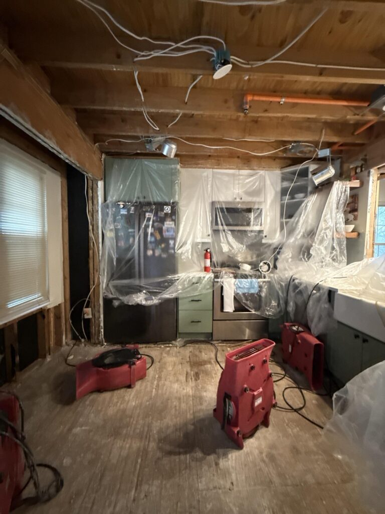 Water damaged kitchen with exposed ceiling, plastic-covered appliances, and red drying fans on the floor.