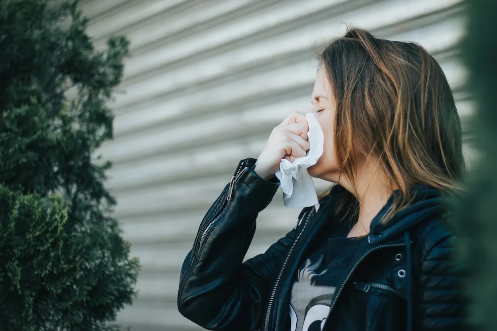 Young woman wearing a black leather jacket with a white tissue in her hand outside – Girl blowing her nose due to allergic reaction or virus infection – Concept image for allergy and having a flu