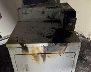 A clothes dryer scorched and blackened by fire, with soot staining the control panel and front — the aftermath of a dryer fire that started in the vent