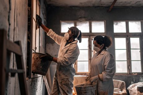 Two workers wearing protective suits, gloves, and face masks clean and repair the interior of a fire-damaged room, using buckets and tools to remove soot and debris from the walls.