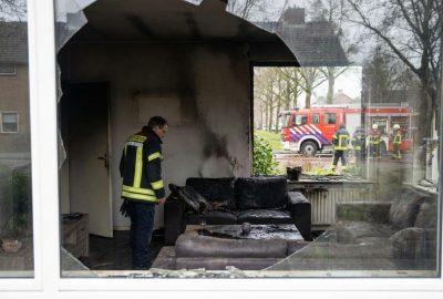 Fire-damaged living room with broken window, charred furniture, and firefighters visible outside during emergency response