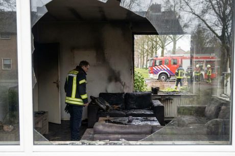 Fire-damaged living room with broken window, charred furniture, and firefighters visible outside during emergency response
