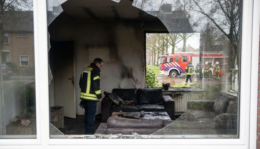 Fire-damaged living room with broken window, charred furniture, and firefighters visible outside during emergency response