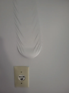 Peeled paint or wallpaper hangs above a broken, discolored electrical outlet.