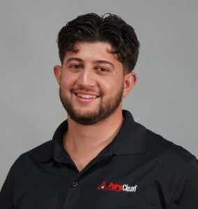 Smiling man with short curly hair wearing a black polo shirt with a logo, set against a gray background.