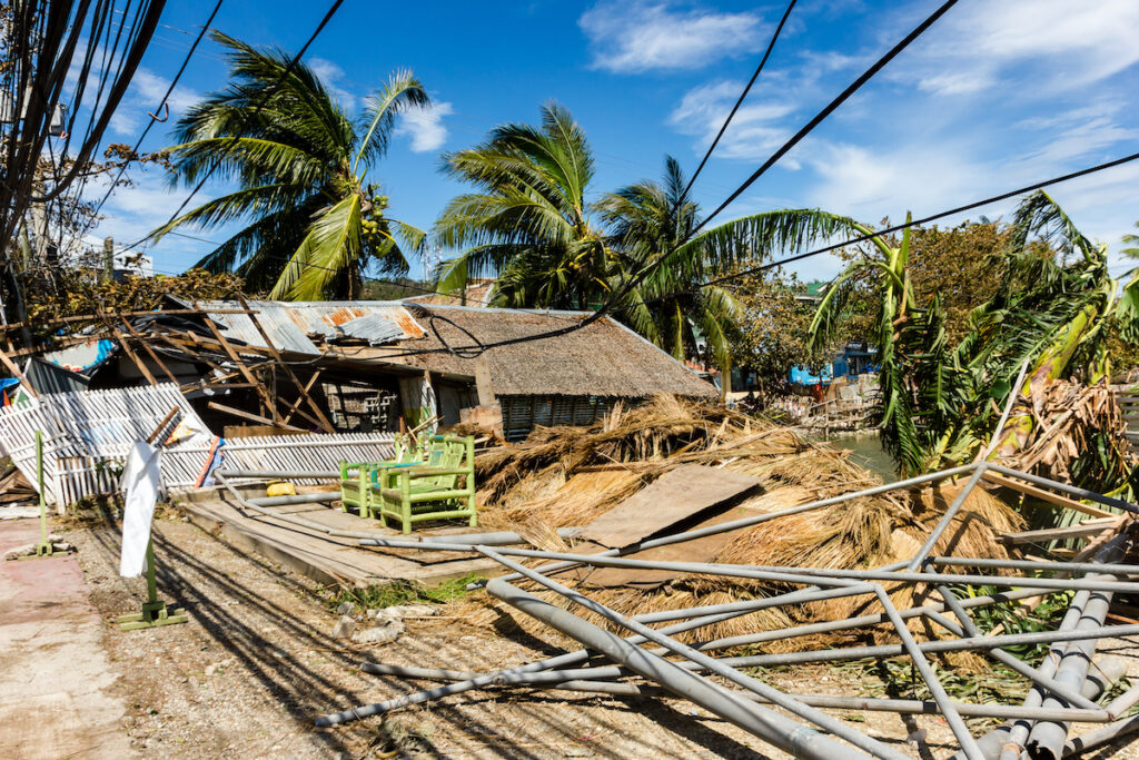 A tropical village shows widespread destruction with damaged homes and scattered debris after a storm.