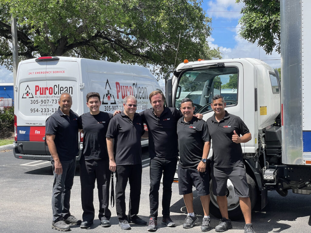 A group of six PuroClean franchise owners in black shirts pose beside their service vehicles in a sunny outdoor setting.