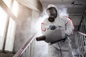 A person in a protective suit uses a fogging machine to disinfect a stairwell filled with mist.