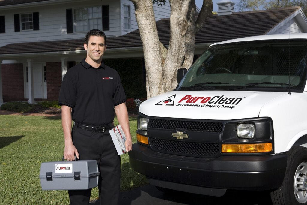 A smiling PuroClean technician holds a toolbox next to a company van in front of a
