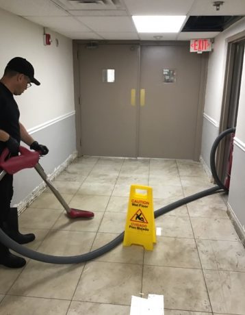 Worker wet-vacuums a tiled hallway floor with a "Caution Wet Floor" sign near