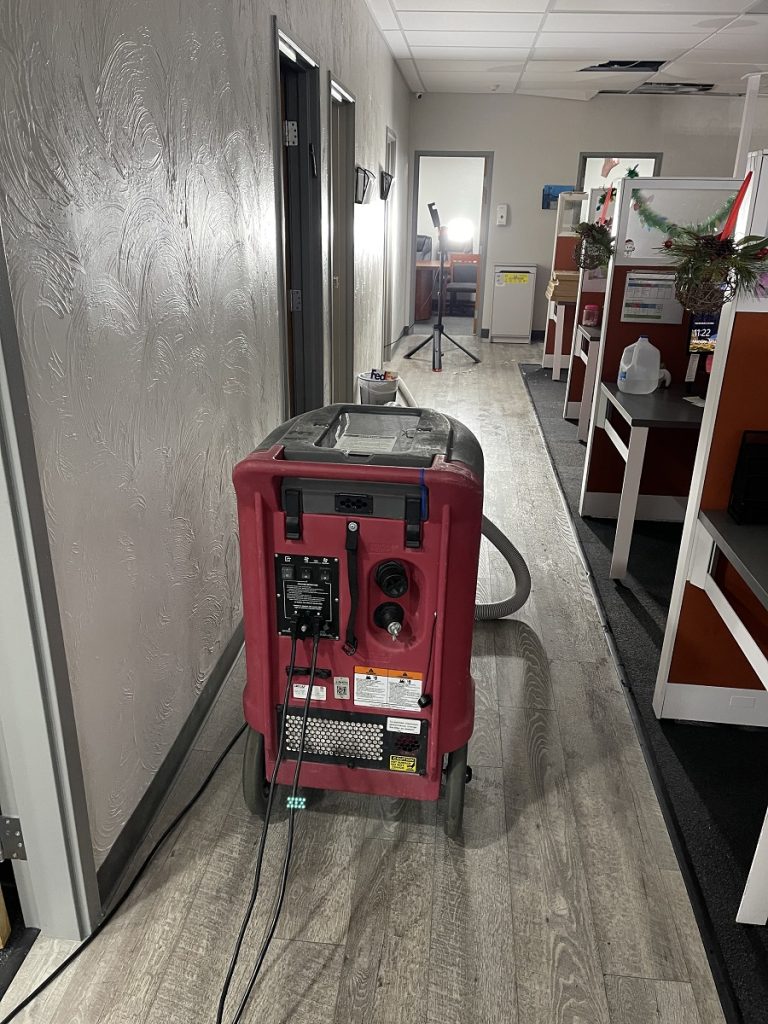 A red industrial machine with hoses and cords sits in an office hallway next to cubicles