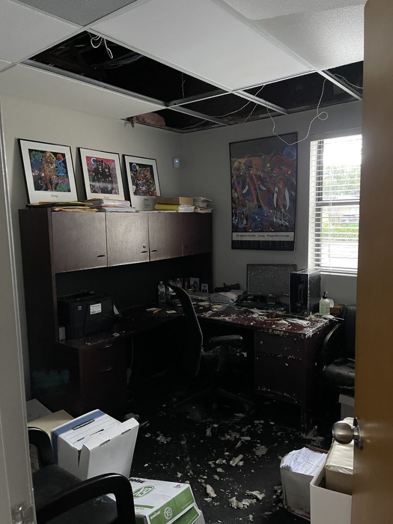 A damaged office interior features missing ceiling tiles, exposed wires, and debris scattered across the floor