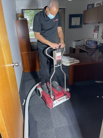 A professional in a mask cleans a dark office carpet with a red PureClean machine. TITLE