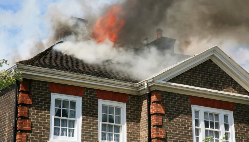 Smoke and flames billow from the roof of a brick house, indicating a fire emergency.