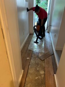 A person uses a shop vacuum to remove water from a partially stripped, wet hallway floor.