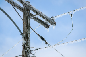 Icicles hang from a wooden utility pole and power lines against a blue sky, illustrating ice storm damage.