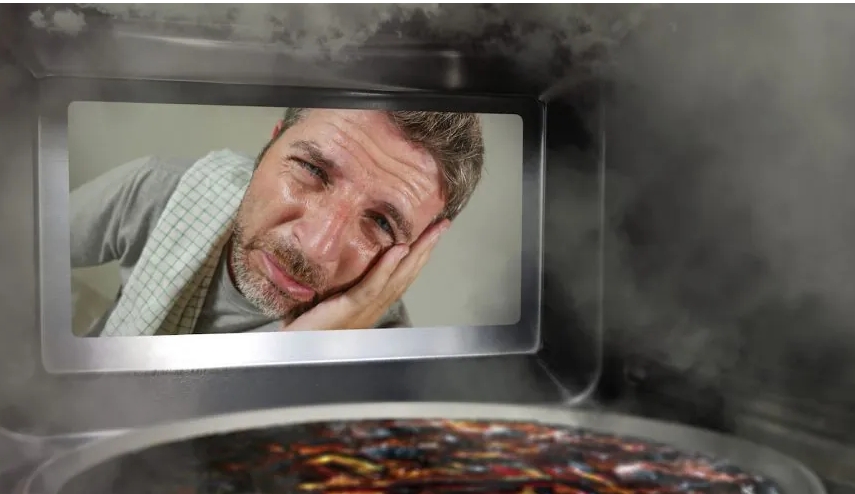 A concerned man looks at a burnt meal inside a smoky microwave, illustrating the aftermath of cooking mishaps.