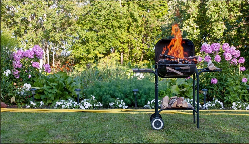 A charcoal grill with flames stands in a garden surrounded by colorful flowers and lush greenery.