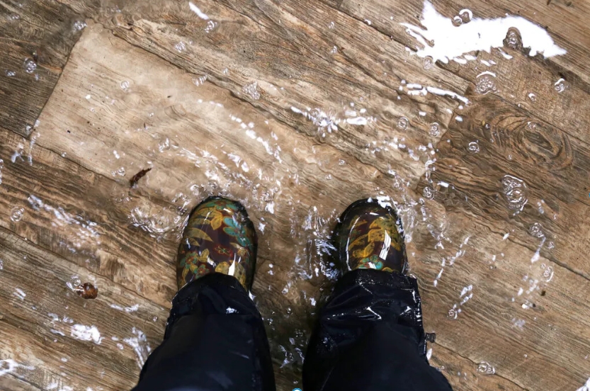Feet in camo boots standing in a flooded basement with water covering a wooden floor.