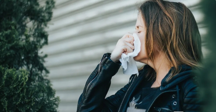 A woman wipes her nose with a tissue, showing signs of allergy discomfort outdoors.