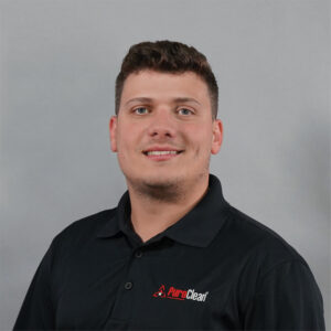 Smiling young man with dark hair wears a black PuroClean polo shirt against a gray background.