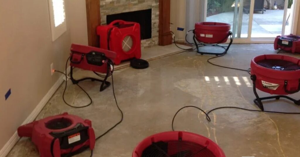 Multiple red air movers and dehumidifiers are actively drying a large room's subfloor.