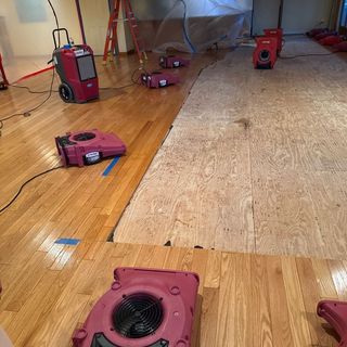 Red drying fans and dehumidifiers are actively drying a room with a partially removed hardwood