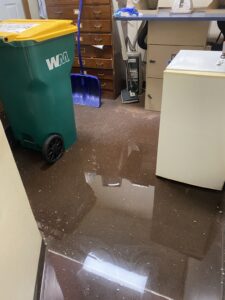 Standing water covers a brown floor in a room with a green recycling bin and wooden cabinets.