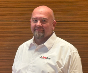 A smiling bald man with a goatee wears a white PureClean shirt against a textured wooden background.