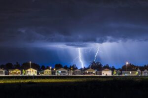 Dramatic lightning strikes illuminate a dark, stormy sky over a line of houses at night.