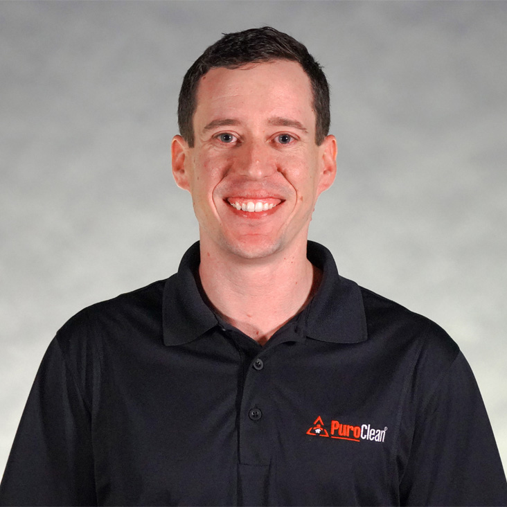 Smiling man in a black polo shirt with a logo, set against a light gray background.