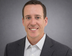 A smiling man in a dark gray suit and white shirt looks directly at the camera. TITLE