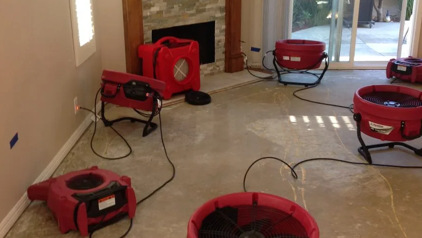 A room undergoing water damage restoration features several red industrial drying fans on the bare floor