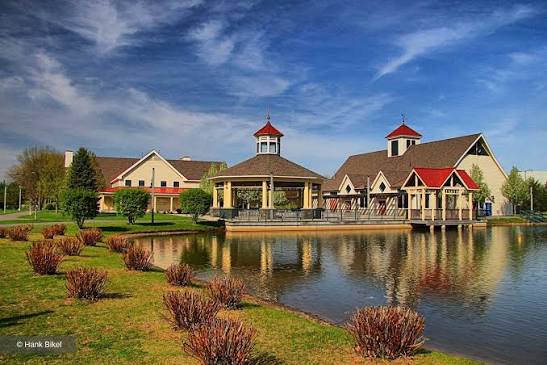 A charming lakeside village features a central gazebo and buildings with brown roofs under a bright blue sky