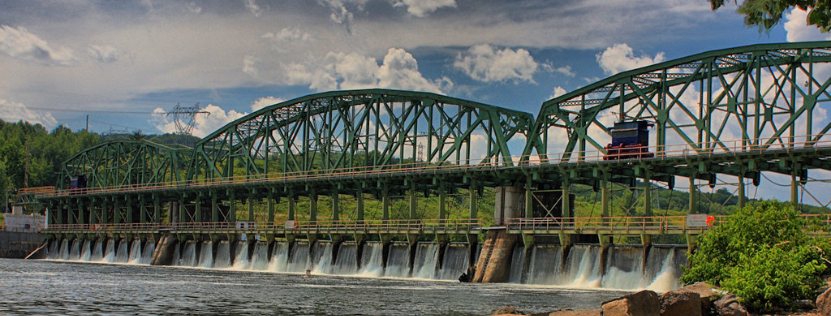 A long green truss bridge with a small train crosses a dam with water spilling over.