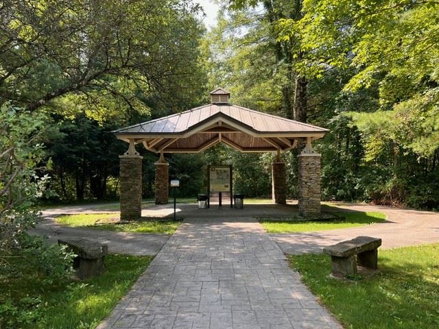 A rustic wooden pavilion with stone pillars sits on a paved path surrounded by lush green
