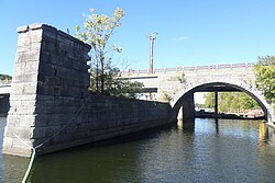 A stone canal wall and an arched bridge span a waterway under a clear blue sky.