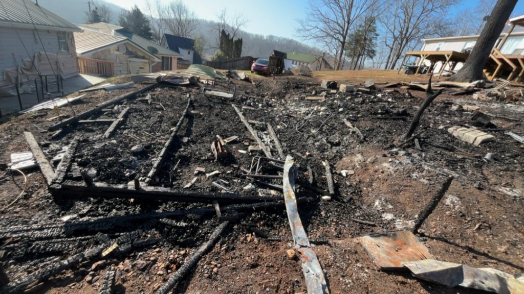 Residential fire mitigation is shown in a heavily destroyed garage structure with collapsed walls, exposed framing, and extensive fire-related debris scattered across the ground. The interior is charred and filled with burned materials, highlighting severe structural loss and smoke damage throughout the building.