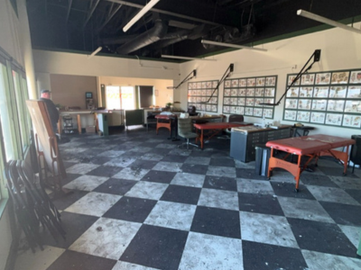 A wide-angle interior shot of a tattoo studio showing extensive smoke and soot damage. The black-and-white checkered floor is heavily marred with dark soot, ash, and scattered debris. Tattoo flash sheets line the white right-hand wall, while the ceiling, exposed ductwork, and upper wall sections are blackened from smoke. Red tattoo beds and equipment are spaced throughout the room.