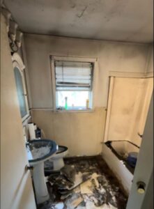 Interior view of a bathroom with extensive damage after a house fire, showing smoke-stained walls and ceiling, water and debris on the floor, and fire damage around the sink and tub.