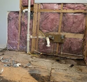 Exposed kitchen wall framing and subfloor showing extensive water damage from a broken pipe leak