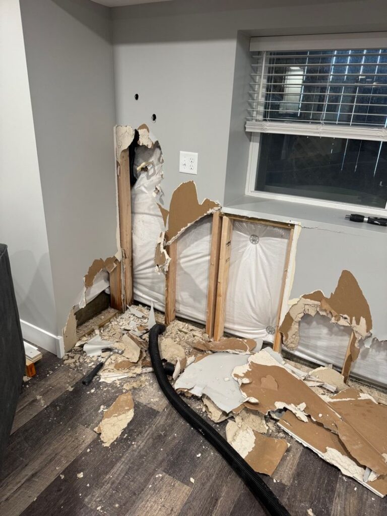A section of a light gray wall under a window has been torn open, revealing wooden wall studs, insulation, and piping, likely to investigate or repair minor leaks. Debris, broken drywall, a hammer, and a black vacuum hose litter the dark wood-look floor below.