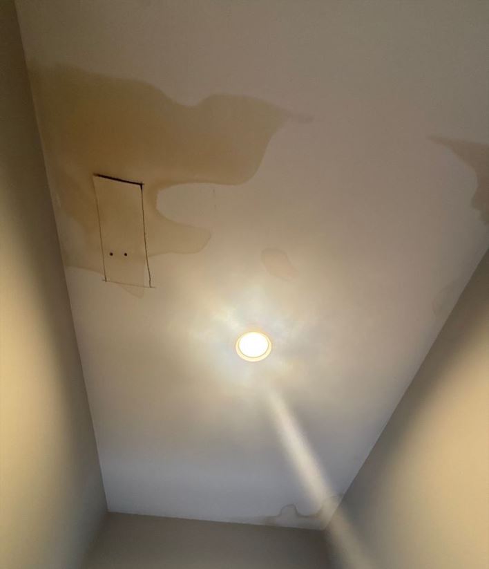 An upward-angled shot of a hallway ceiling showing a severe water stain spreading across the surface. The large, yellowish-brown water stain has distinct, irregular rings and covers a significant portion of the upper left side of the ceiling, surrounding a small rectangular access panel. A glowing, recessed circular light fixture in the center casts a bright glare downward, while smaller, fainter patches of water stain discoloration are visible along the upper right edges and corners of the ceiling.