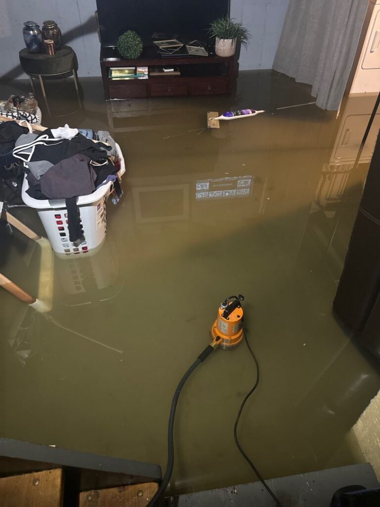 A heavily flooded finished basement with several inches of standing, murky, greenish-brown water covering the entire floor. In the foreground, an orange submersible utility pump is submerged in the water to drain the basement, connected to a black discharge hose and power cord. In the background, a white plastic laundry basket filled with clothes, a wooden TV stand, a small side table, and other household belongings are partially submerged in the basement floodwater.