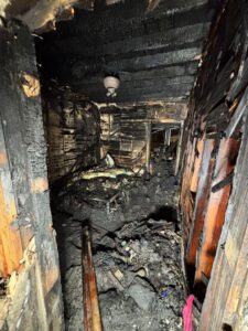Completely charred room showing severe damage from an electrical fire with melted ceiling fan fixture and exposed wooden framing.