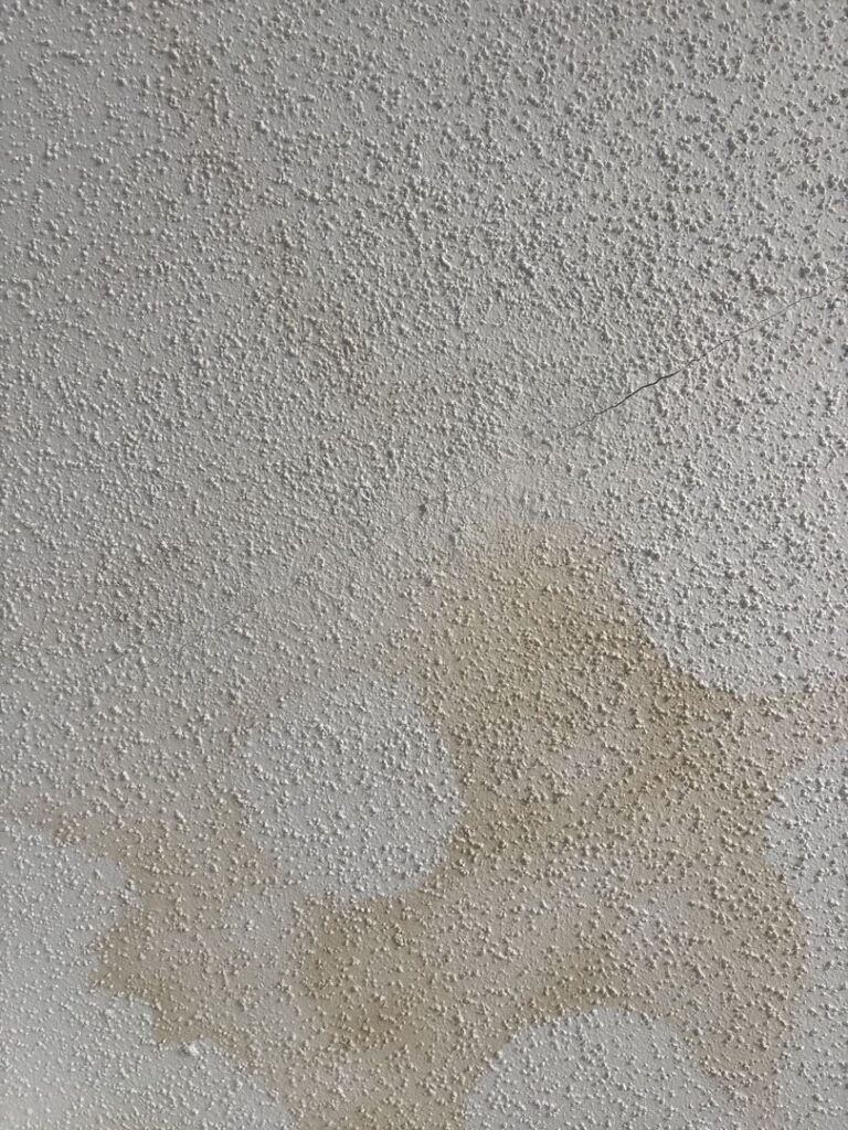 A close-up view of a textured popcorn ceiling showing clear signs of moisture damage. A large, irregular, light-yellowish water stain spreads across the bumpy, white stippled surface. A thin, diagonal hairline crack cuts through the texture, running directly above the stained area.