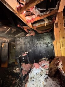 Severe house fire damage showing charred walls, exposed wooden ceiling framing, and collapsed pink insulation inside a home.